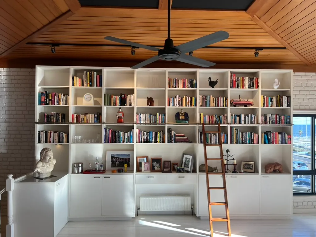 Wall of bookshelves with books, art pieces, ladder, and framed photos, set against a white brick wall with a ceiling fan above.
