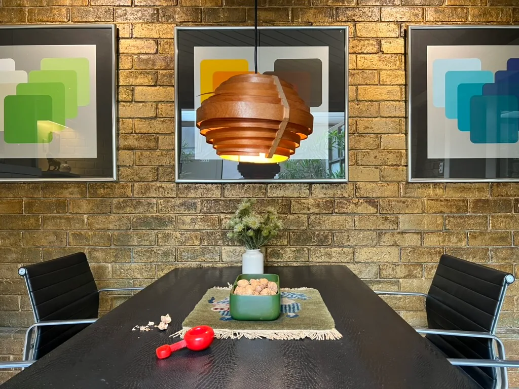 Dining area with a modern pendant lamp, artwork on a brick wall, black chairs, and a table with decor and a red rattle.