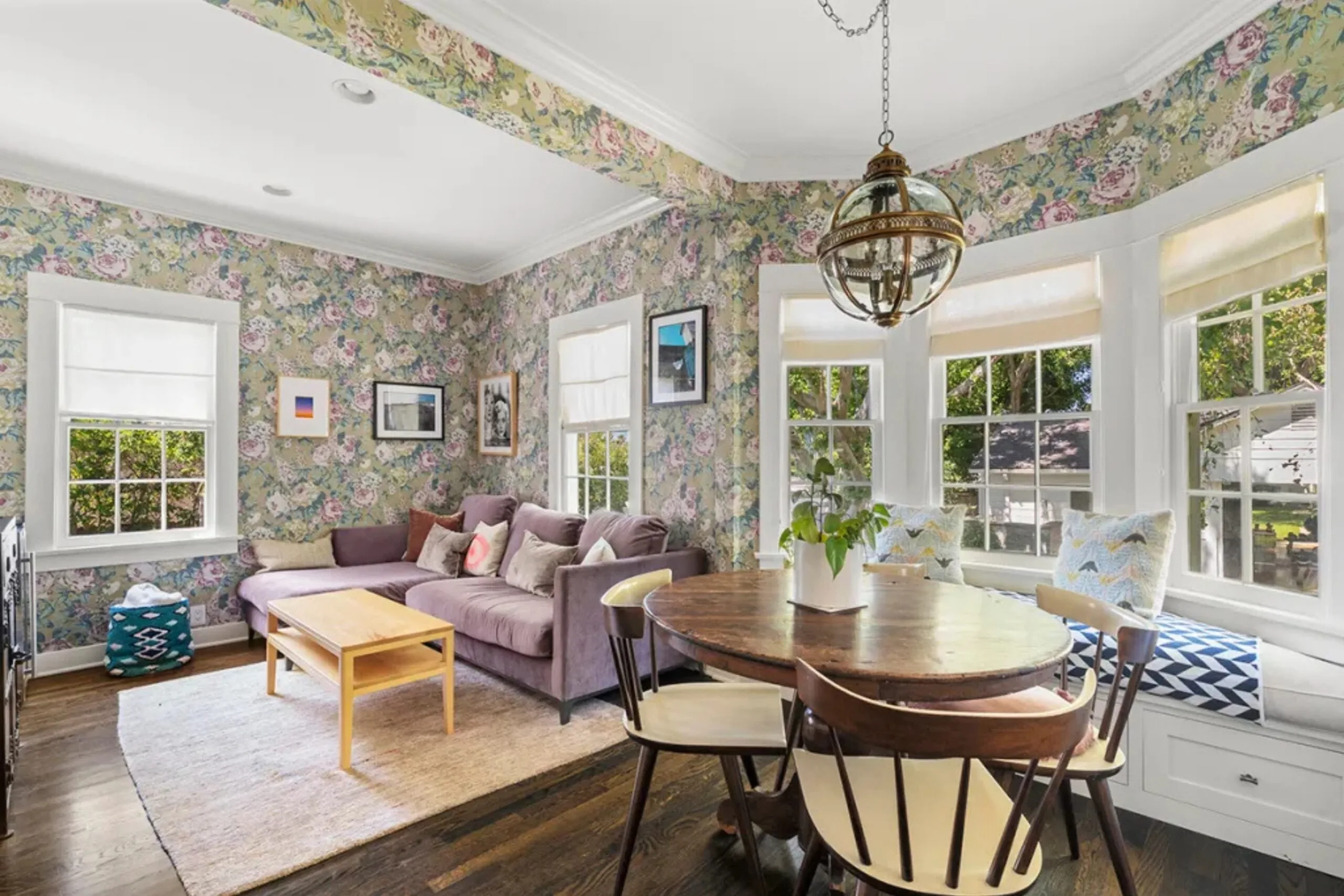 Living room with floral wallpaper, purple sofa, wooden dining table, window seat, and large bright windows.