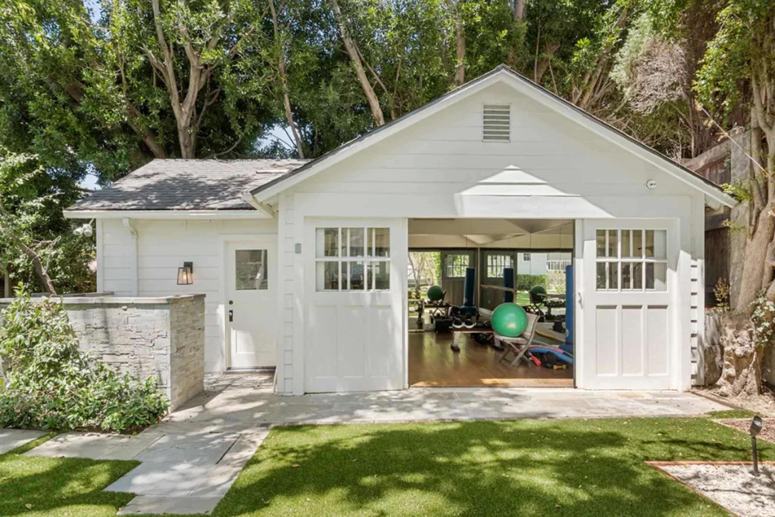 White garage with open doors revealing a home gym inside, surrounded by lush green trees and grass.