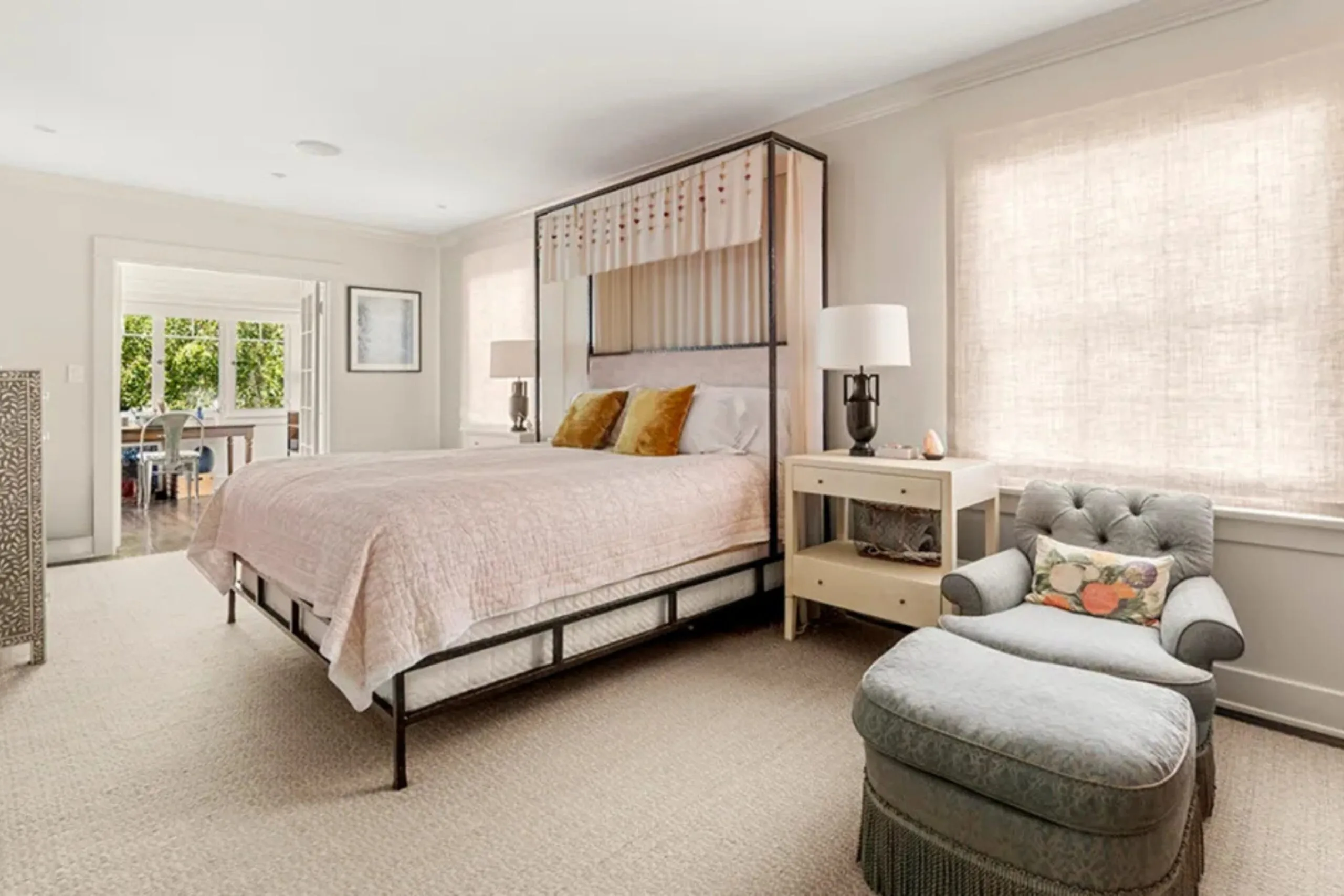 Bedroom with a canopy bed, light pink bedding, side tables with lamps, and a green upholstered chair and ottoman.