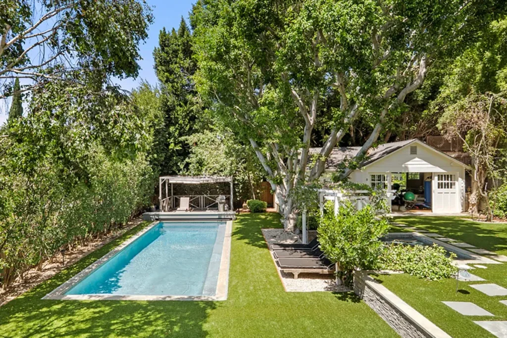Backyard with a rectangular pool, lush greenery, trees, and a small white pool house.