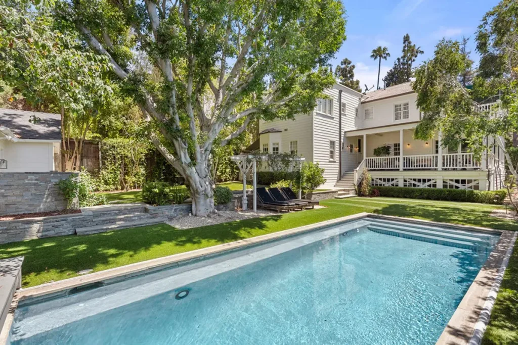 Backyard view of a house with a pool, large tree, and lawn, surrounded by greenery and white siding on a sunny day.