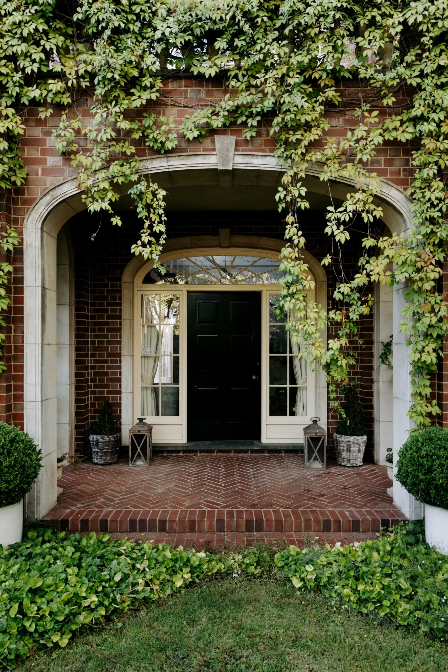 Brick archway with ivy and a black door, flanked by lanterns and potted plants on a red brick patio.