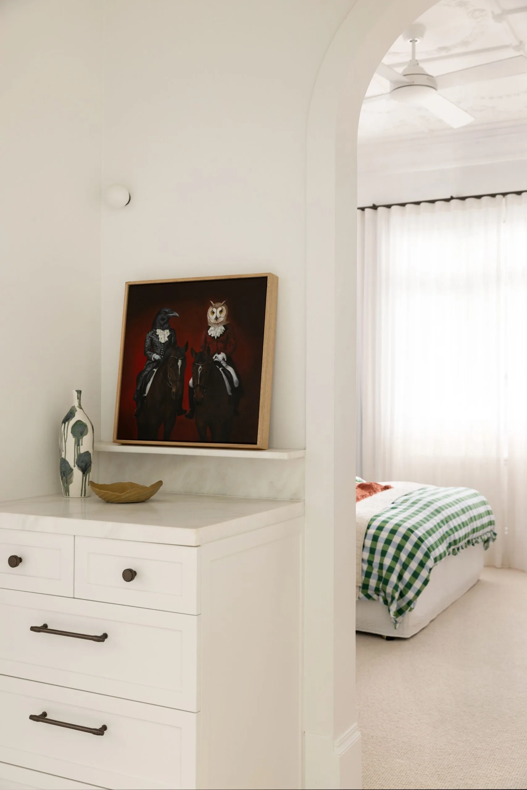 White room with a dresser, a painting of a crow and owl on horseback, a vase, and a glimpse of a bedroom with a patterned bedspread.