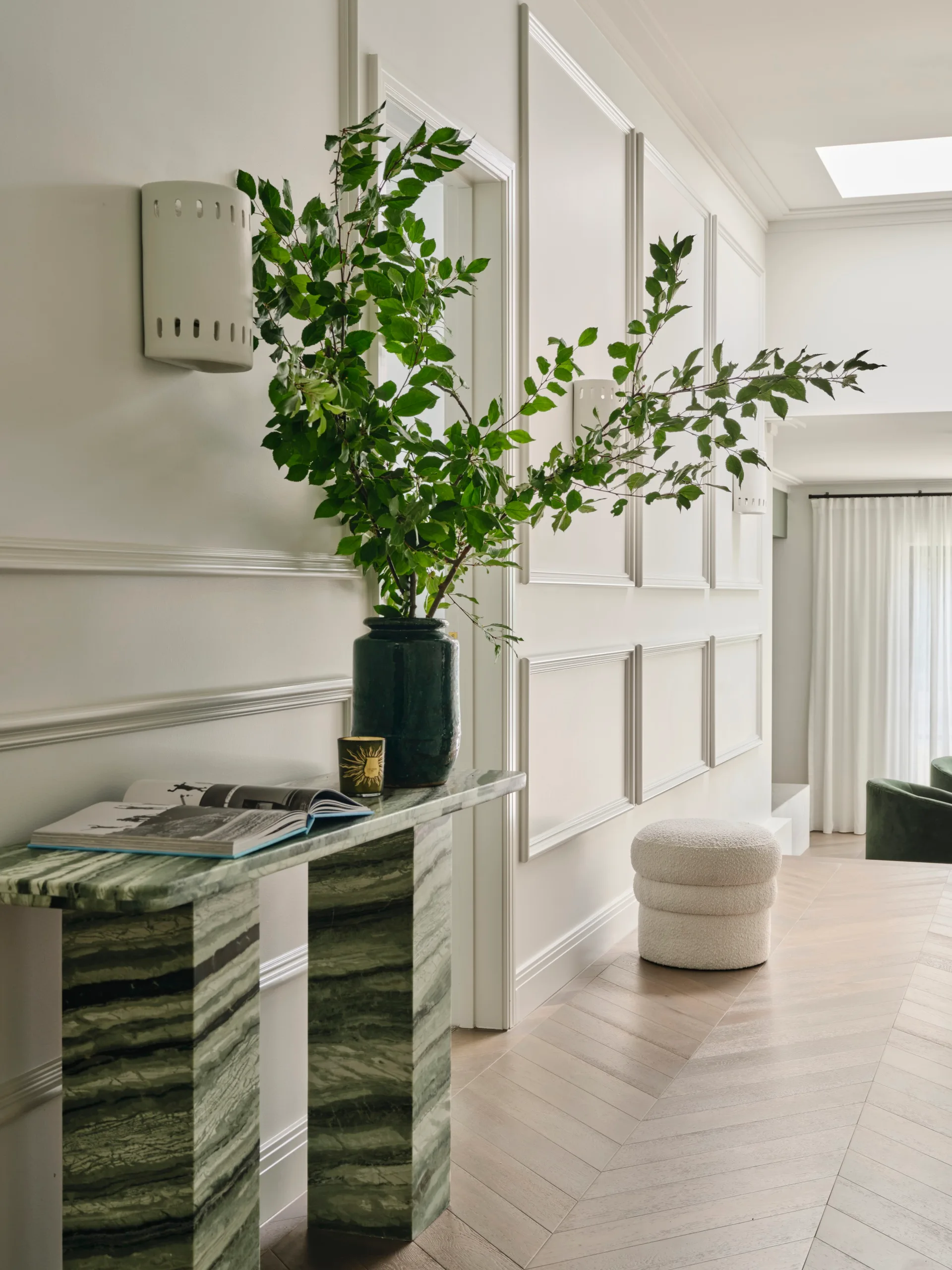 Elegant hallway with a green marble console table, leafy plant, and book, against paneled white walls.