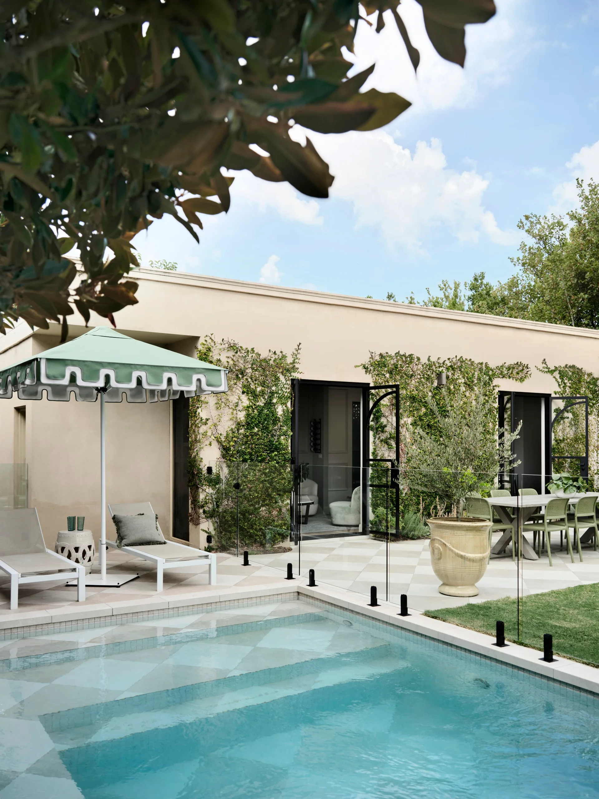 Outdoor pool with glass fence, lounge chairs under green umbrella, and dining table beside a beige house with greenery.