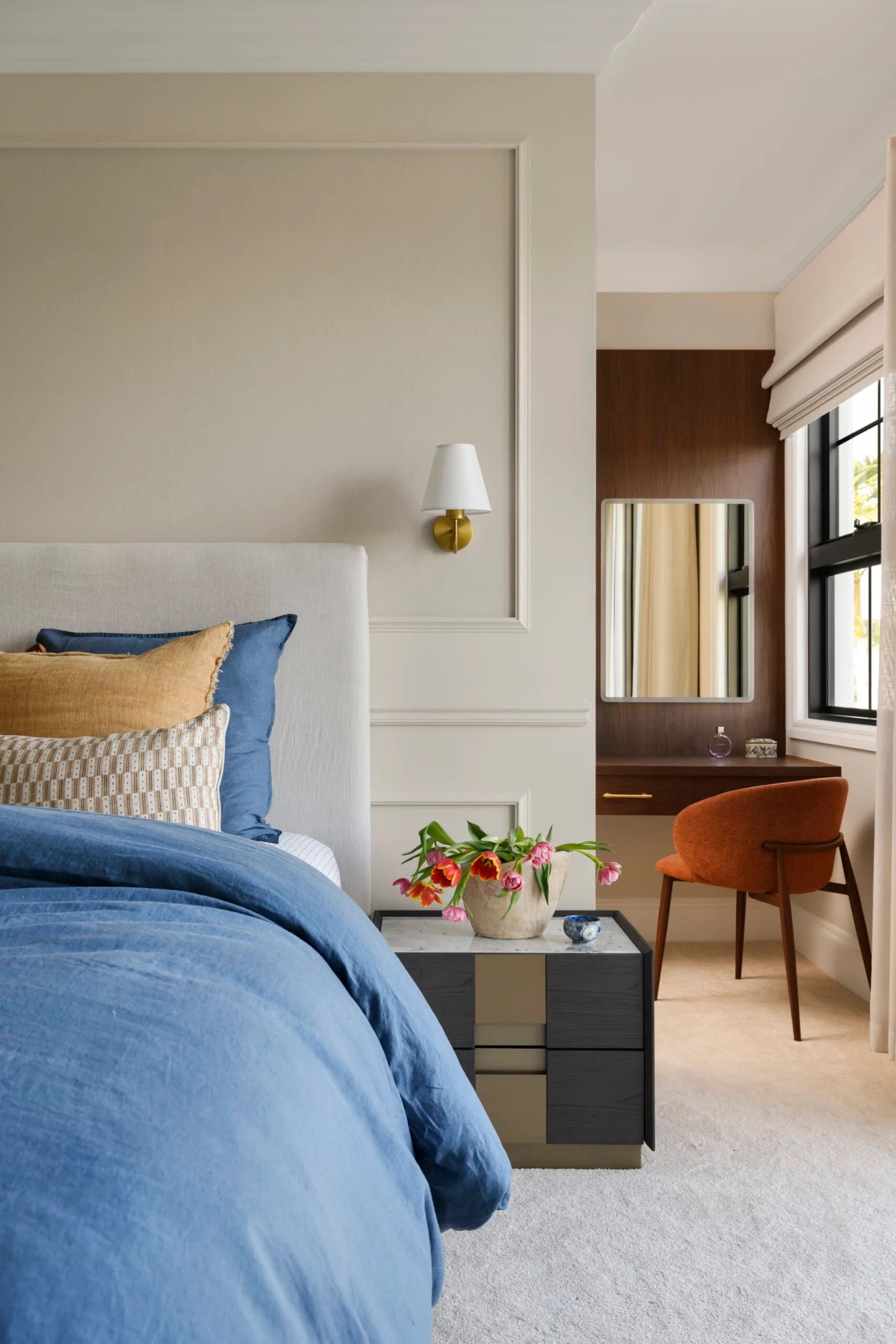 Bedroom with a blue bedspread, accented pillows, a side table with flowers, and a small desk with an orange chair.