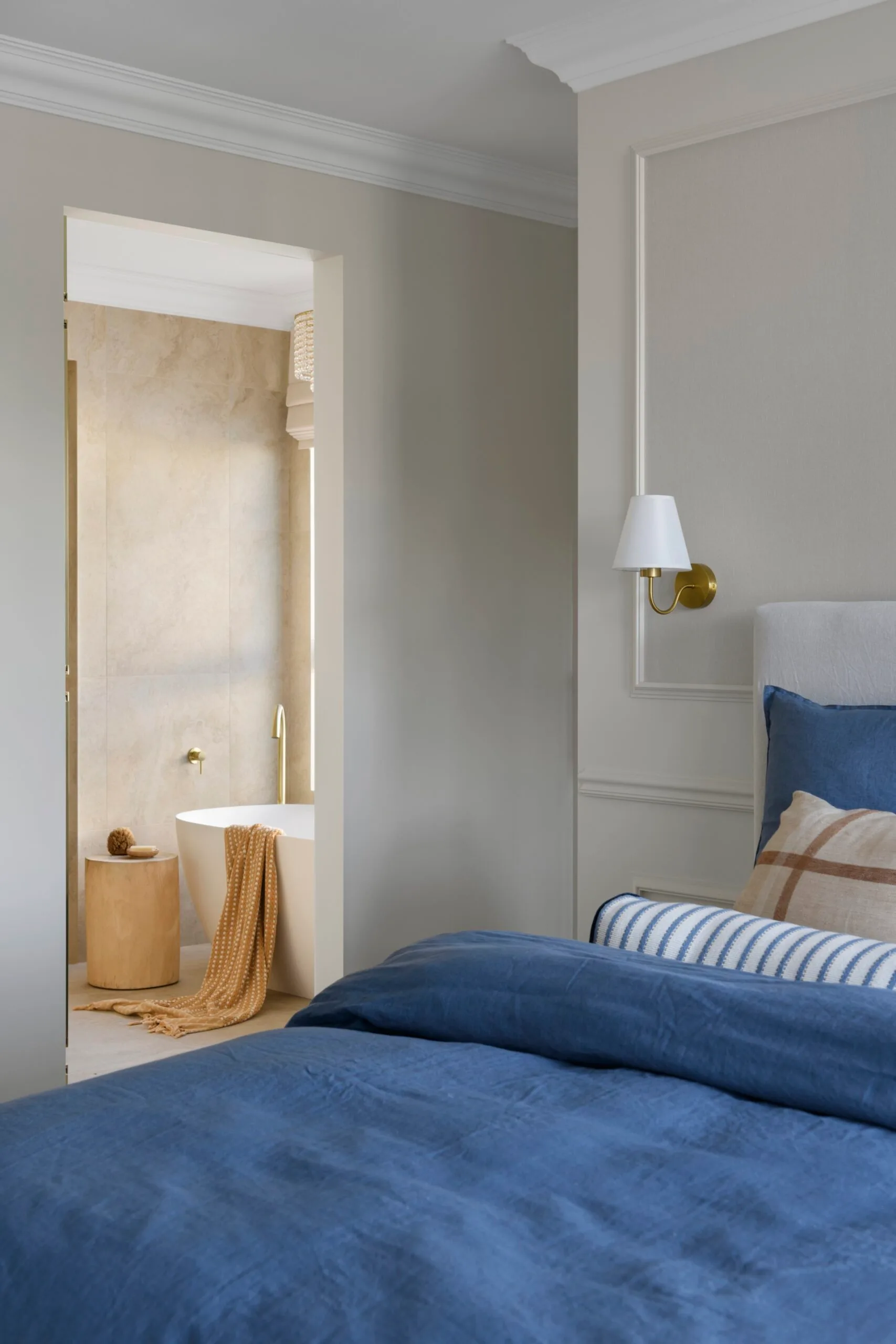 Bedroom with a blue bedspread, pillows, wall lamp, and adjacent bathroom with a freestanding tub and wooden side table.
