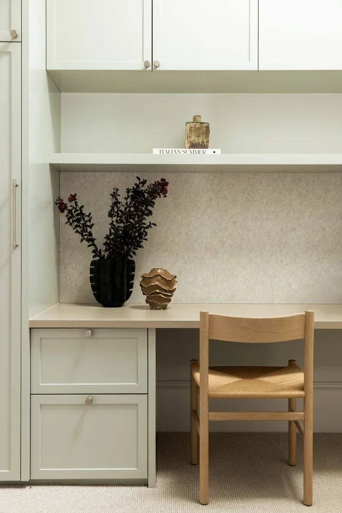 Minimalist desk setup with a wooden chair, decorative vase, and dark plant on a light green cabinet. Book titled "Italian Summer" on shelf.