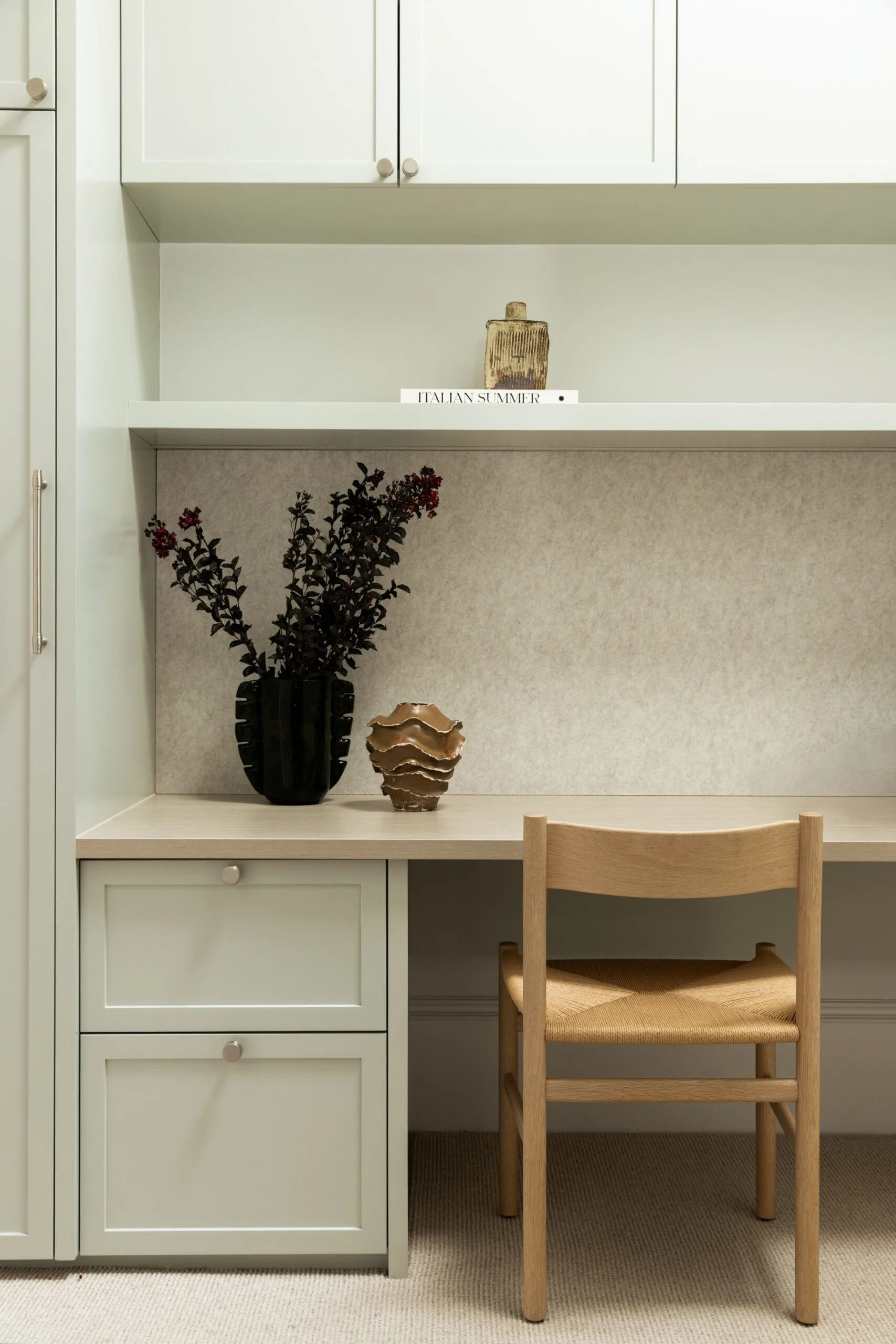 Minimalist desk setup with a wooden chair, decorative vase, and dark plant on a light green cabinet. Book titled "Italian Summer" on shelf.