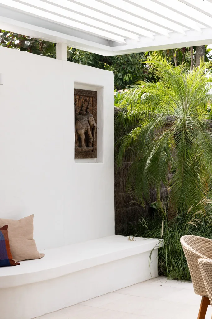 Outdoor seating area with white bench, decorative cushions, elephant wall carving, and tropical plants under a pergola.