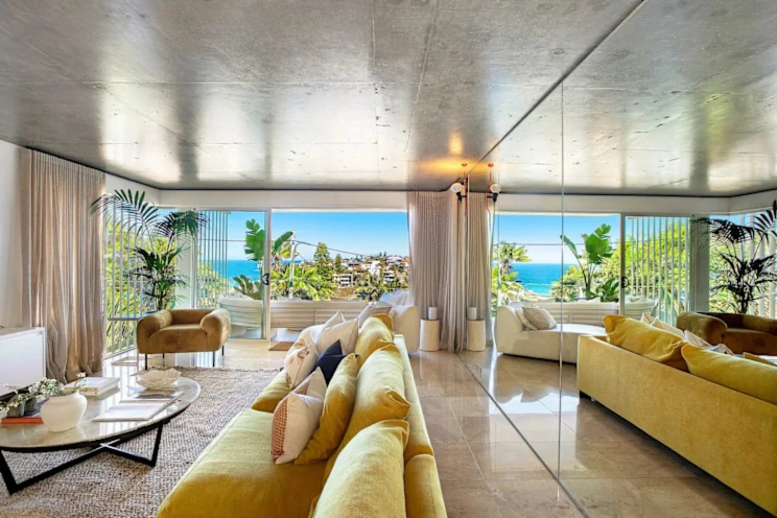 Bright living room with yellow sofas, glass walls, and ocean view from a large window. Nicole O'Neil's new home