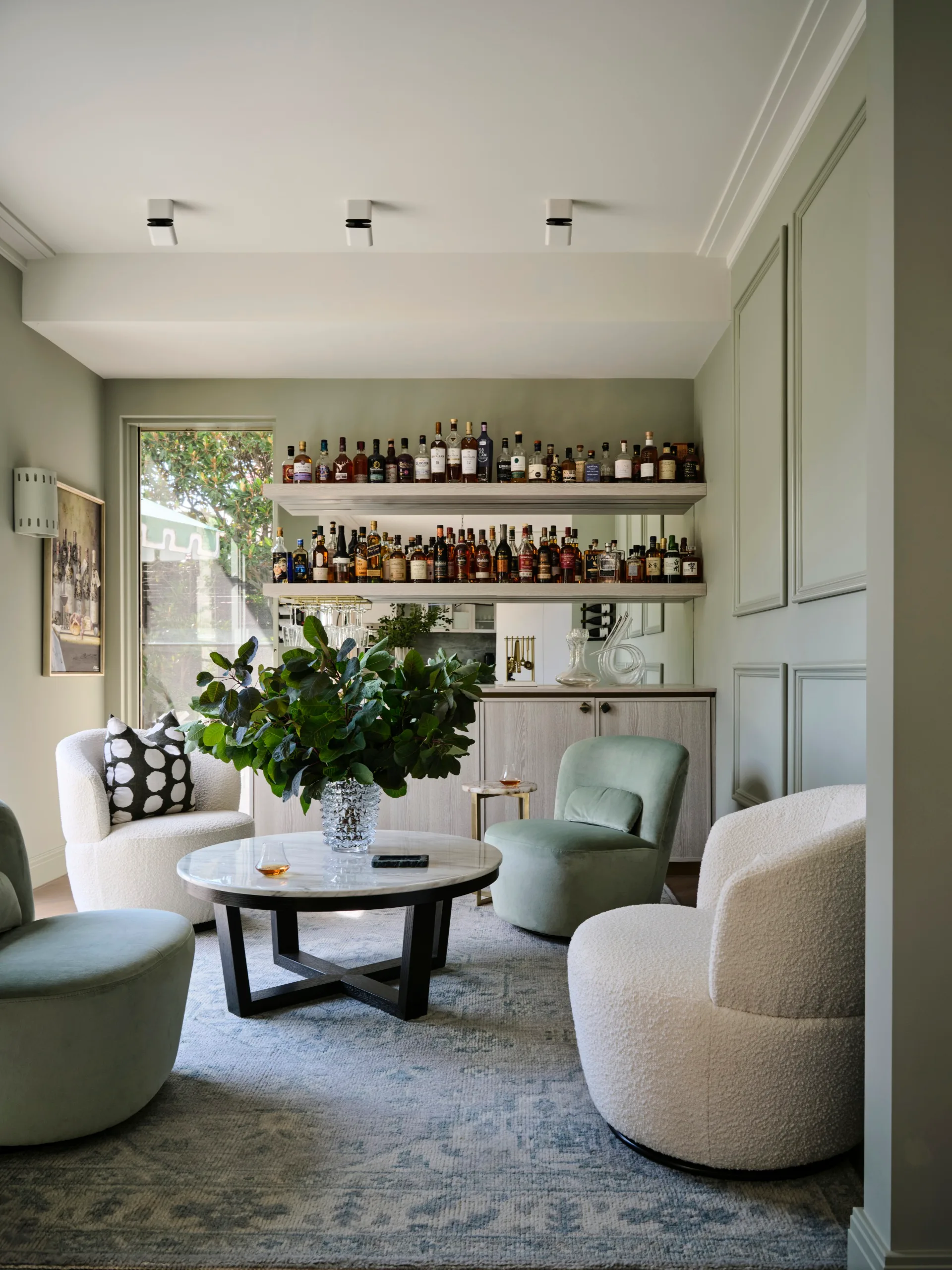 Cozy living room with a bar, plush armchairs, a round marble table, and a shelf of assorted liquor bottles.