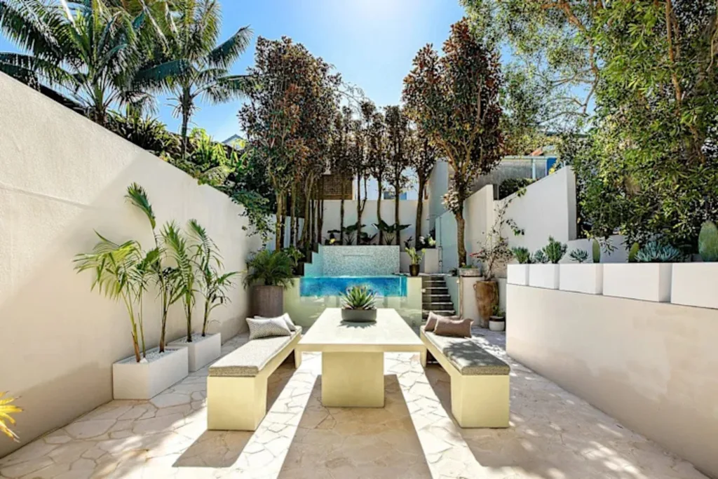 Modern outdoor patio with benches, a central table, wall planters, trees, and a small water feature under bright sunlight. Nicole O'Neil's new home