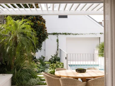 Outdoor patio with a round wooden table and chairs, lush greenery, and a view of a pool in a modern backyard setting.