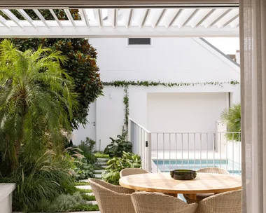 Outdoor patio with a round wooden table and chairs, lush greenery, and a view of a pool in a modern backyard setting.