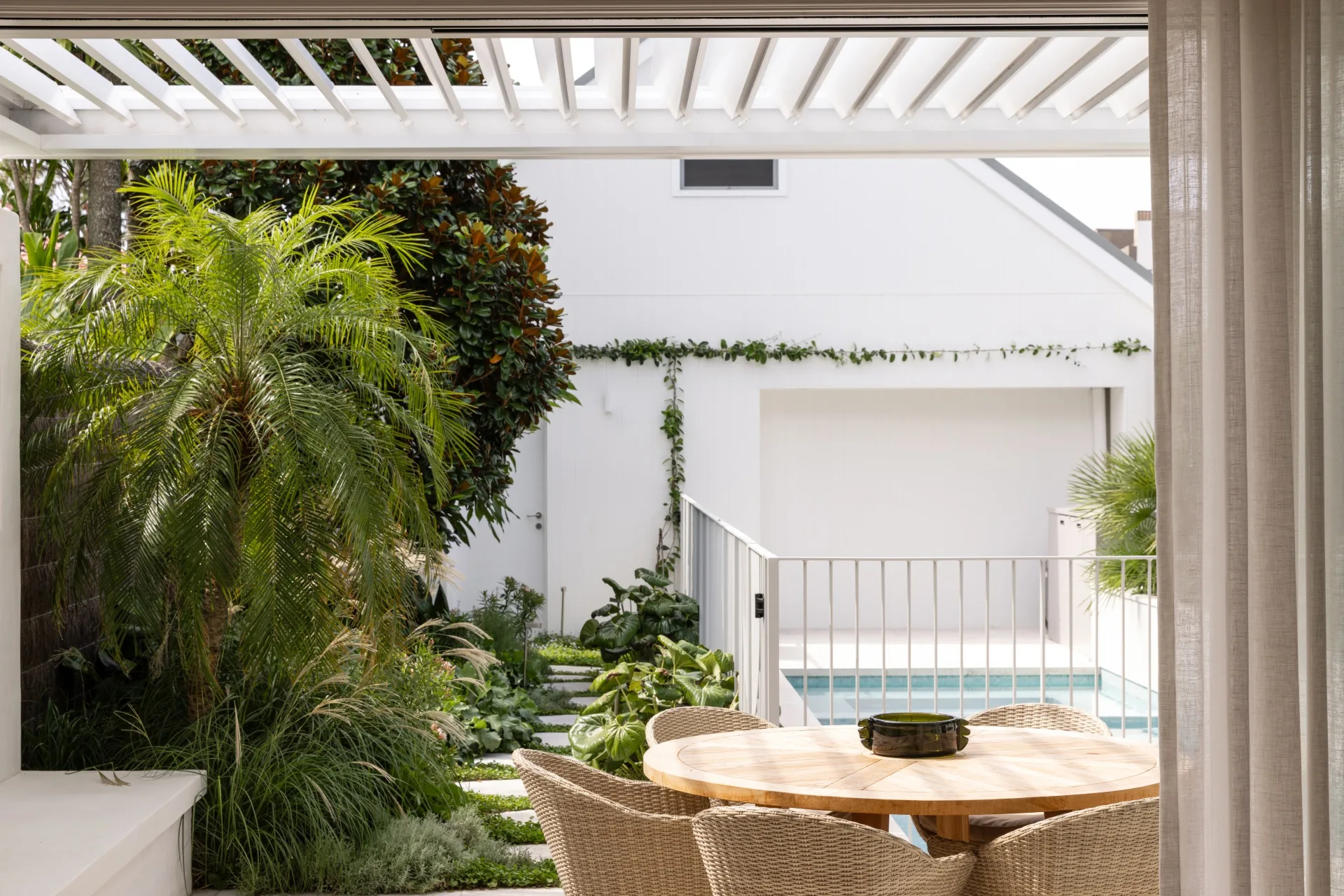 Outdoor patio with a round wooden table and chairs, lush greenery, and a view of a pool in a modern backyard setting.