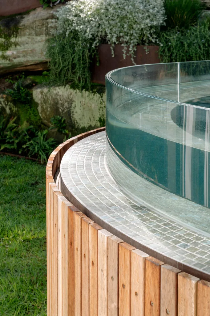 Modern glass-walled swimming pool with wooden and tile accents in a lush garden setting.