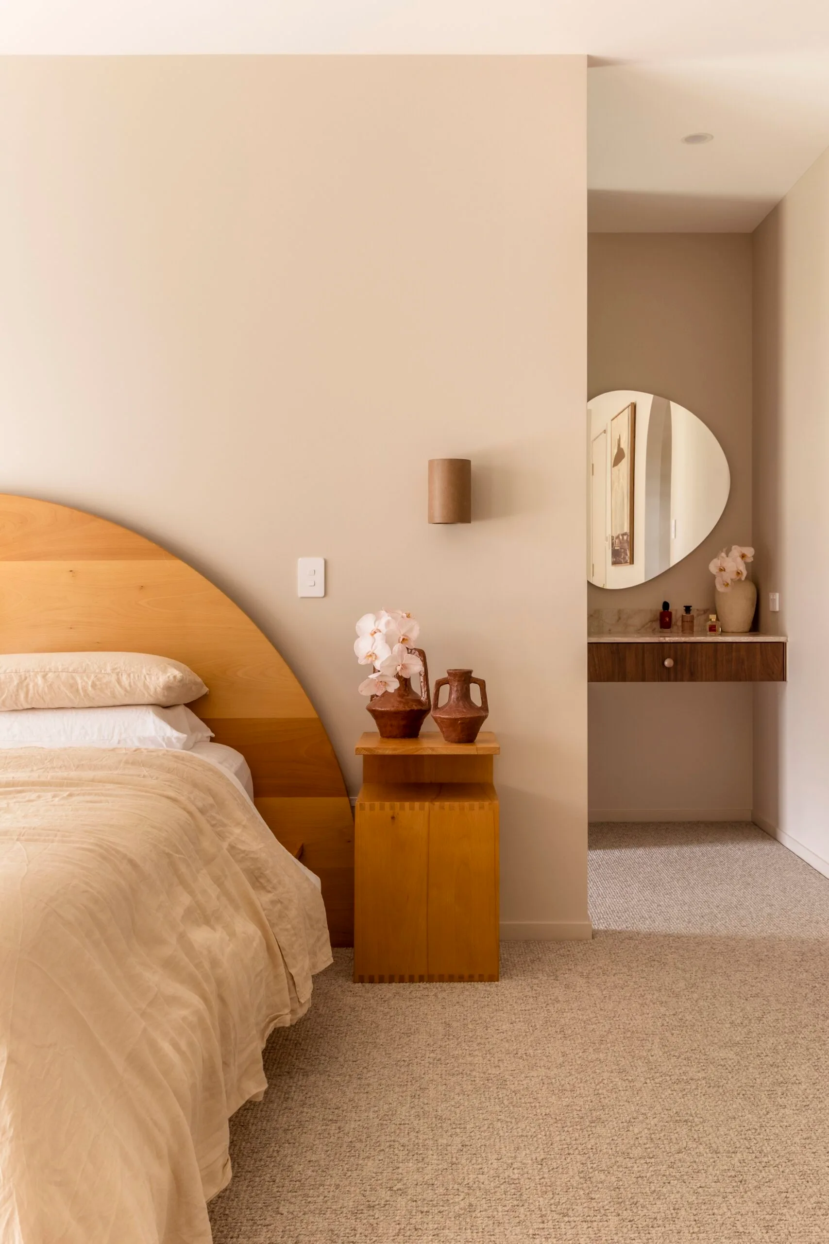 Cozy bedroom with wooden bed and bedside table, beige bedding, pottery decor, and a round mirror in the background.
