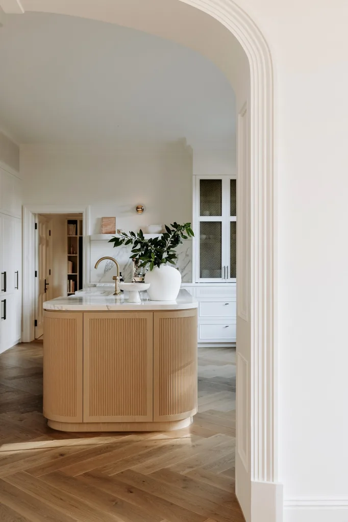 Modern kitchen with wooden island, white cabinets, and a plant in a white vase, viewed through an archway.