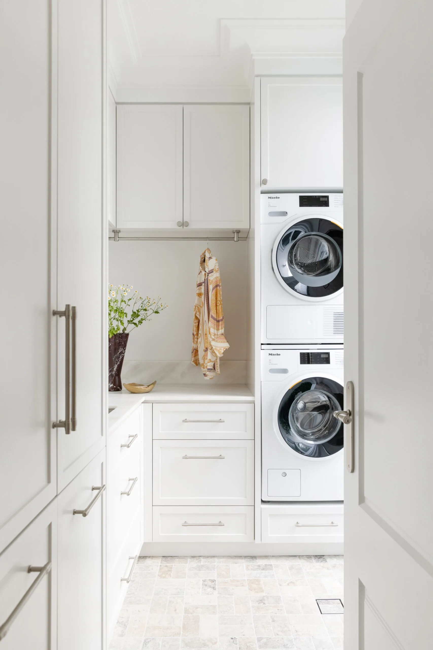 Laundry room with white cabinets, stacked washer and dryer, hanging towel, and a vase with flowers on the counter.