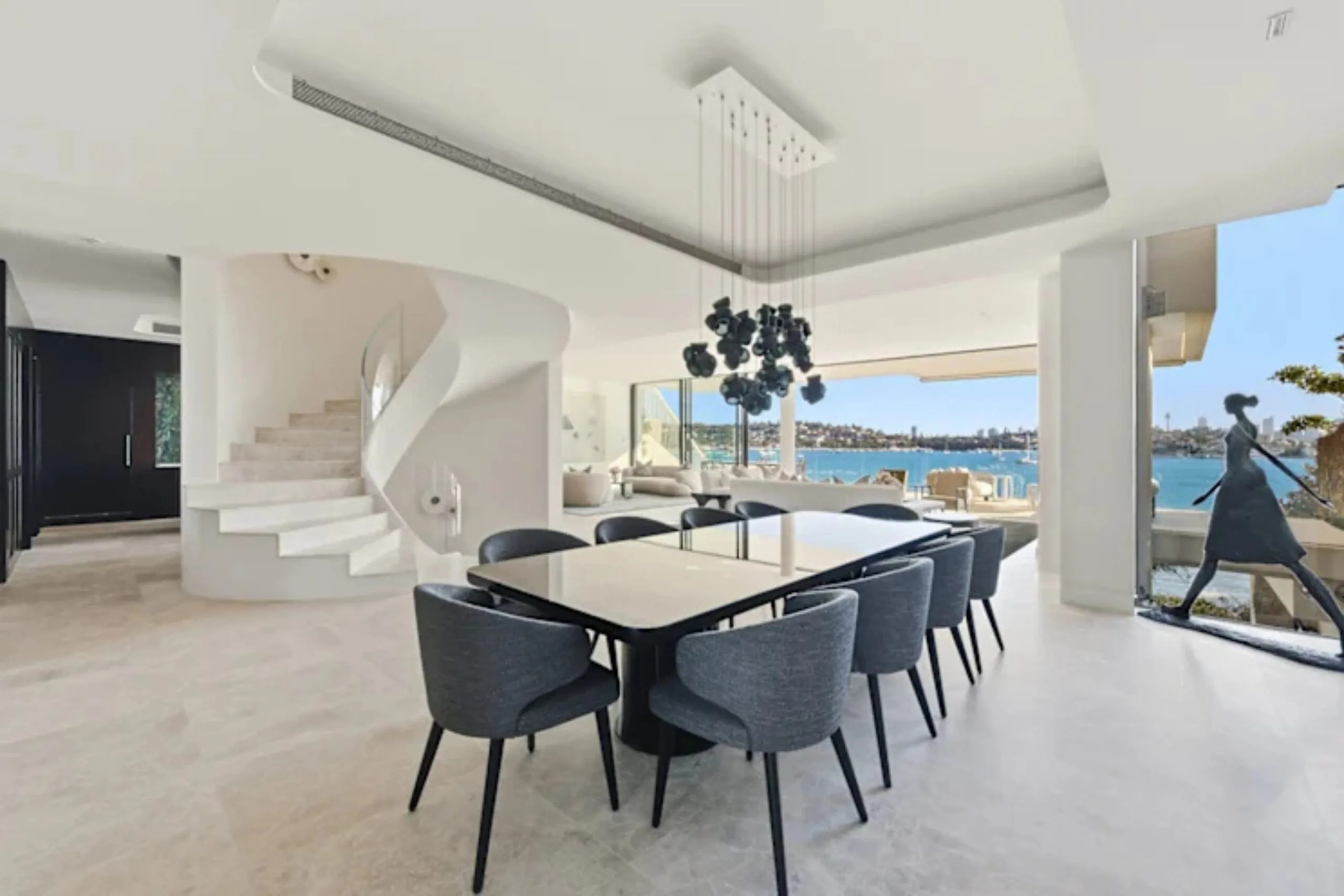 Modern dining room with a large table, spiral staircase, and ocean view through floor-to-ceiling windows. Nicole O'Neil's new home