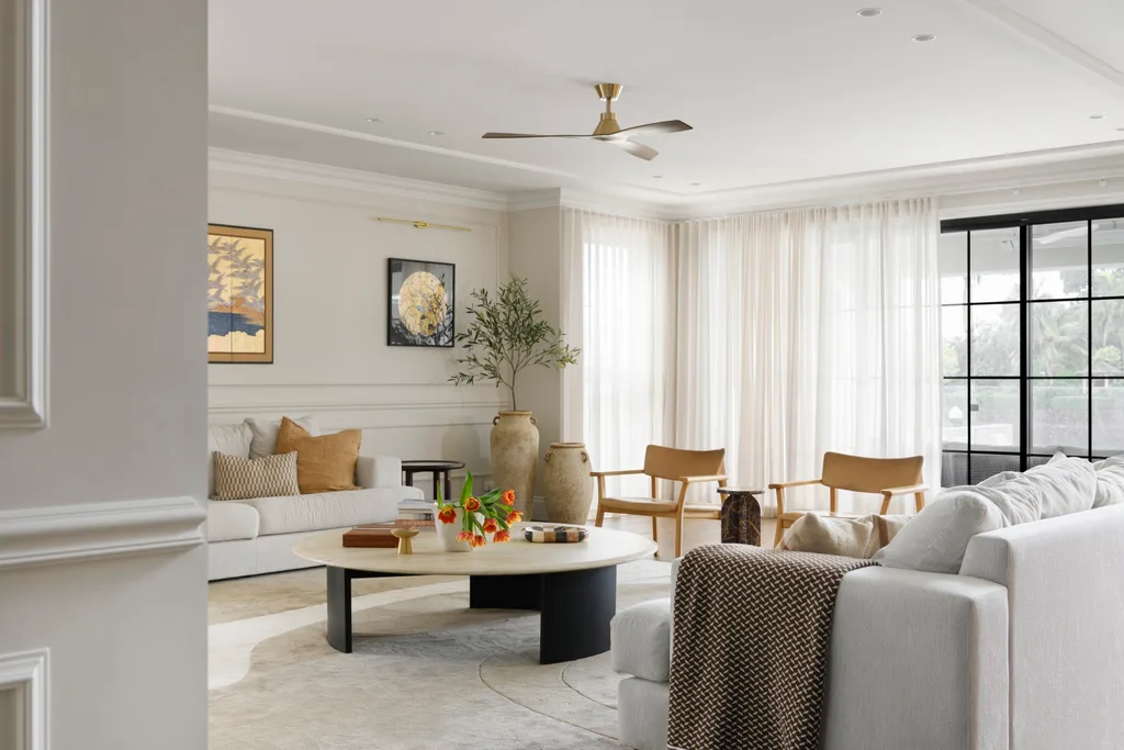 Luxurious living room with neutral tones, sofa, armchairs, a large round coffee table, and soft lighting from sheer curtains.