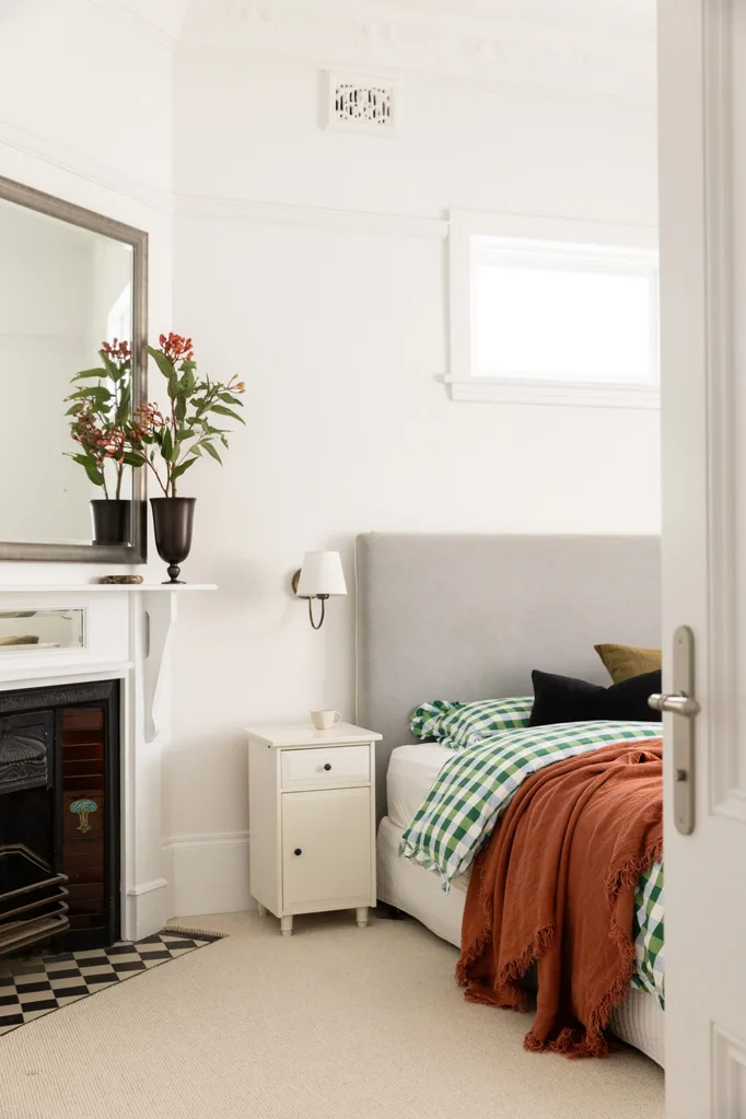 Cozy bedroom with a gray headboard, orange throw, checkered bedding, and a fireplace with a mirror and vase on the mantel.