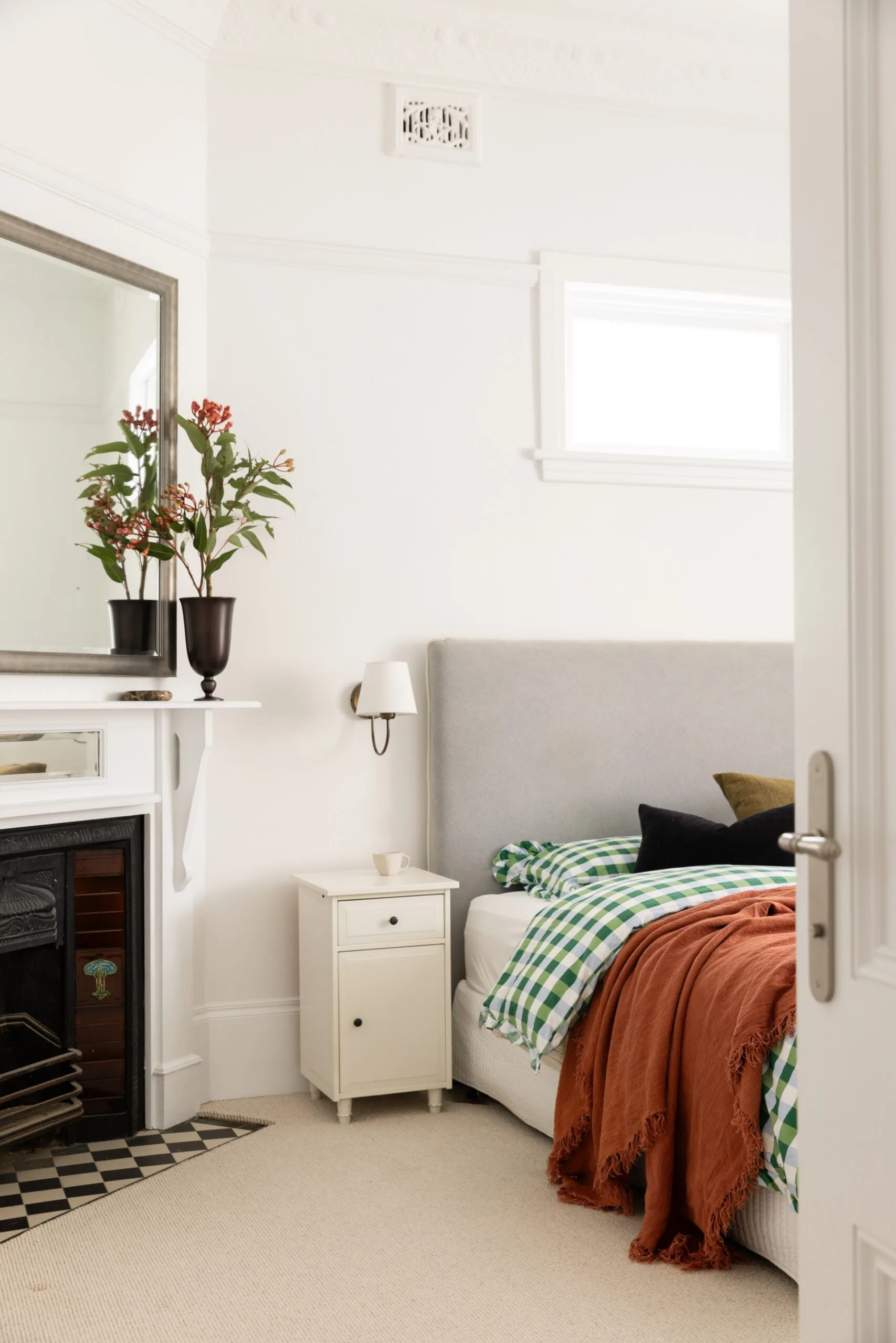 Cozy bedroom with a gray headboard, orange throw, checkered bedding, and a fireplace with a mirror and vase on the mantel.