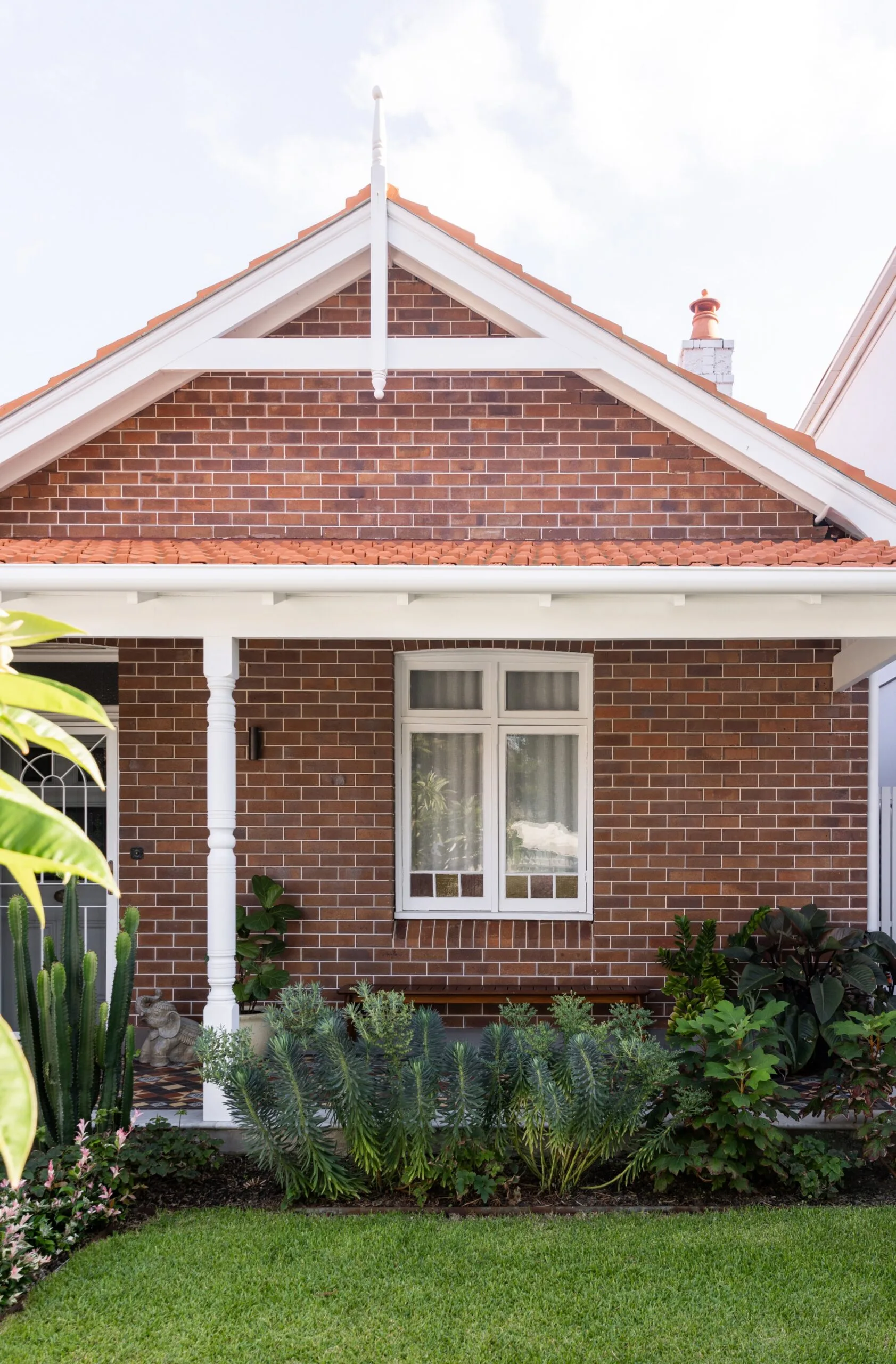 Red brick house with white trim, a front window, and garden shrubs; pitched roof on a sunny day.