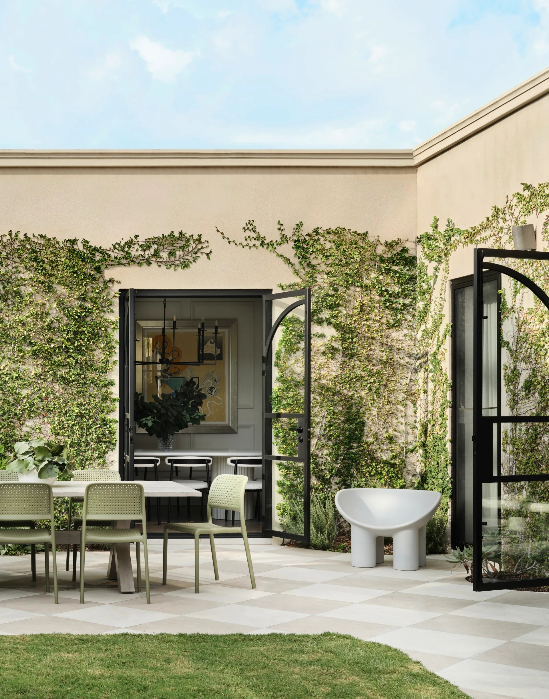Outdoor patio with green chairs, table, white chair, open glass doors, and walls covered in green ivy.