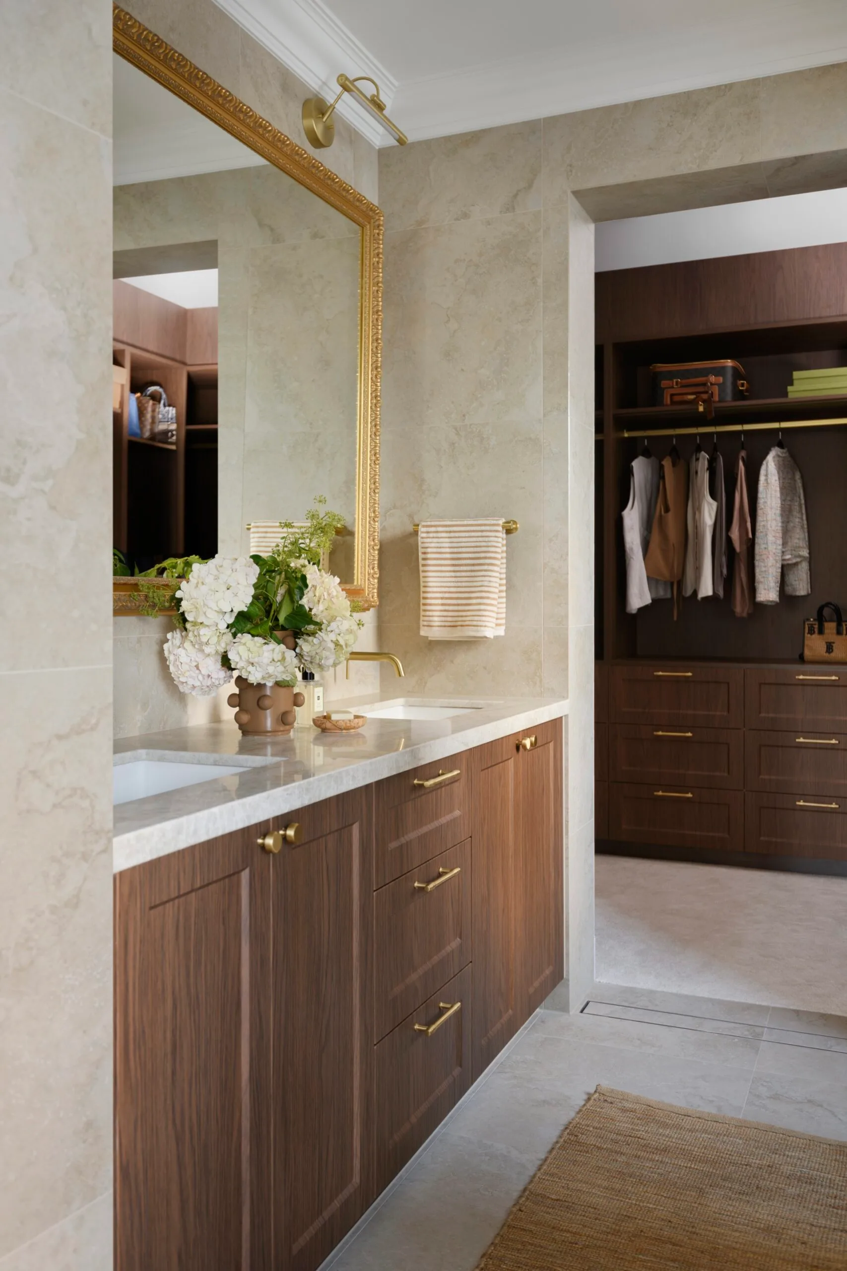 Luxurious bathroom with a wooden vanity, large mirror, brass fixtures, and a vase of white flowers, leading to a closet.
