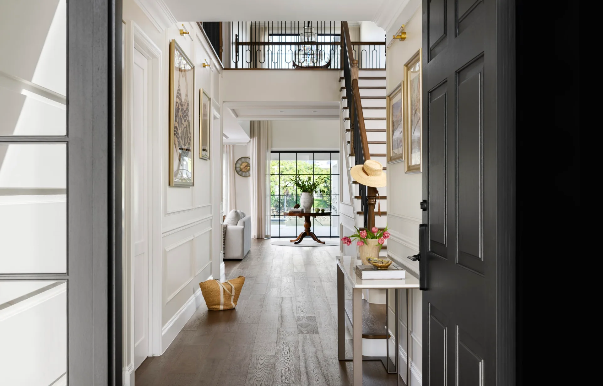 Elegant hallway with dark door, staircase, artwork, a straw hat on the banister, flowers on a table, and a sunny view.