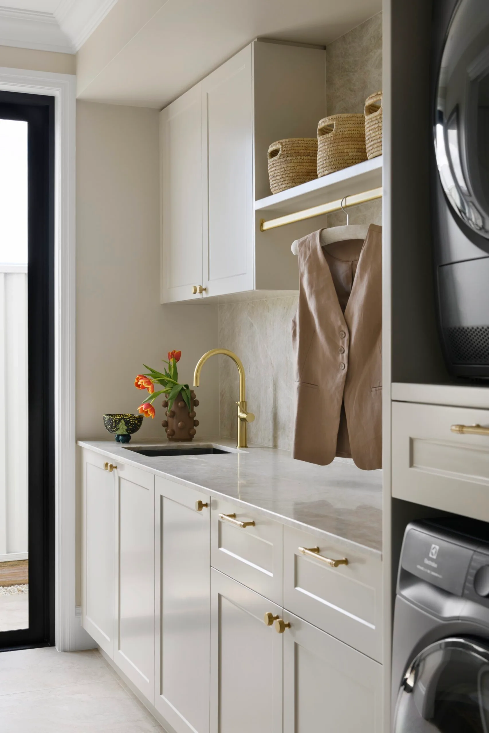 Laundry room with beige cabinets, gold fixtures, hanging vest, and a vase of tulips on the countertop.