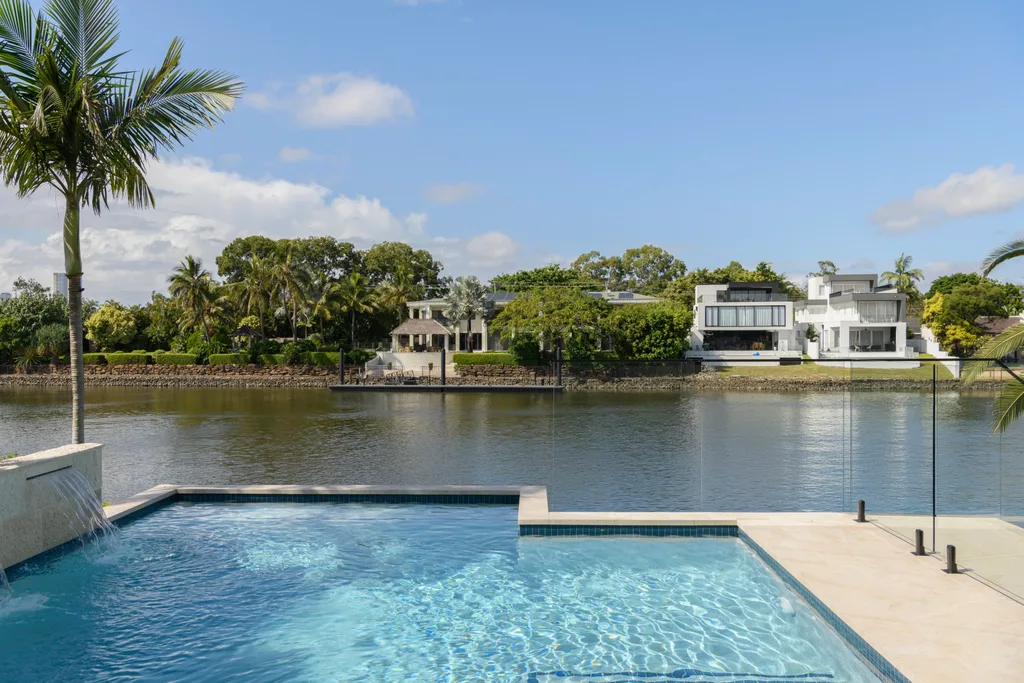 Outdoor pool with water feature overlooking a serene river and luxury houses surrounded by lush trees.