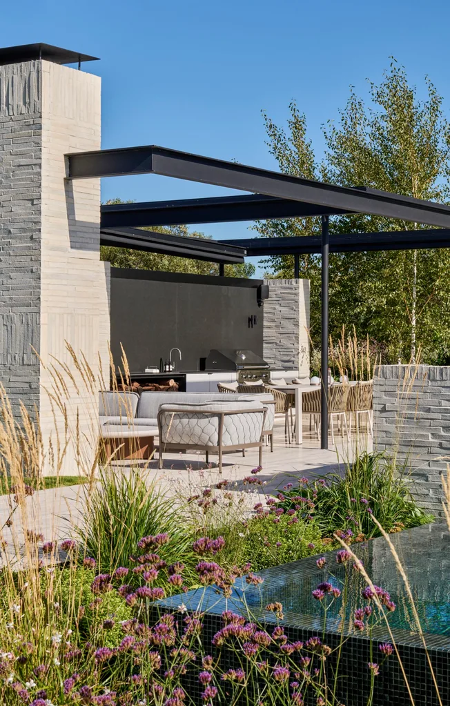 Outdoor patio with modern furniture, pergola, and grill; surrounded by greenery and tall ornamental grasses. garden
