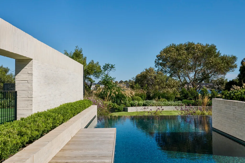 Modern backyard with a large, clear pool, stone walls, lush greenery, and bright blue sky.