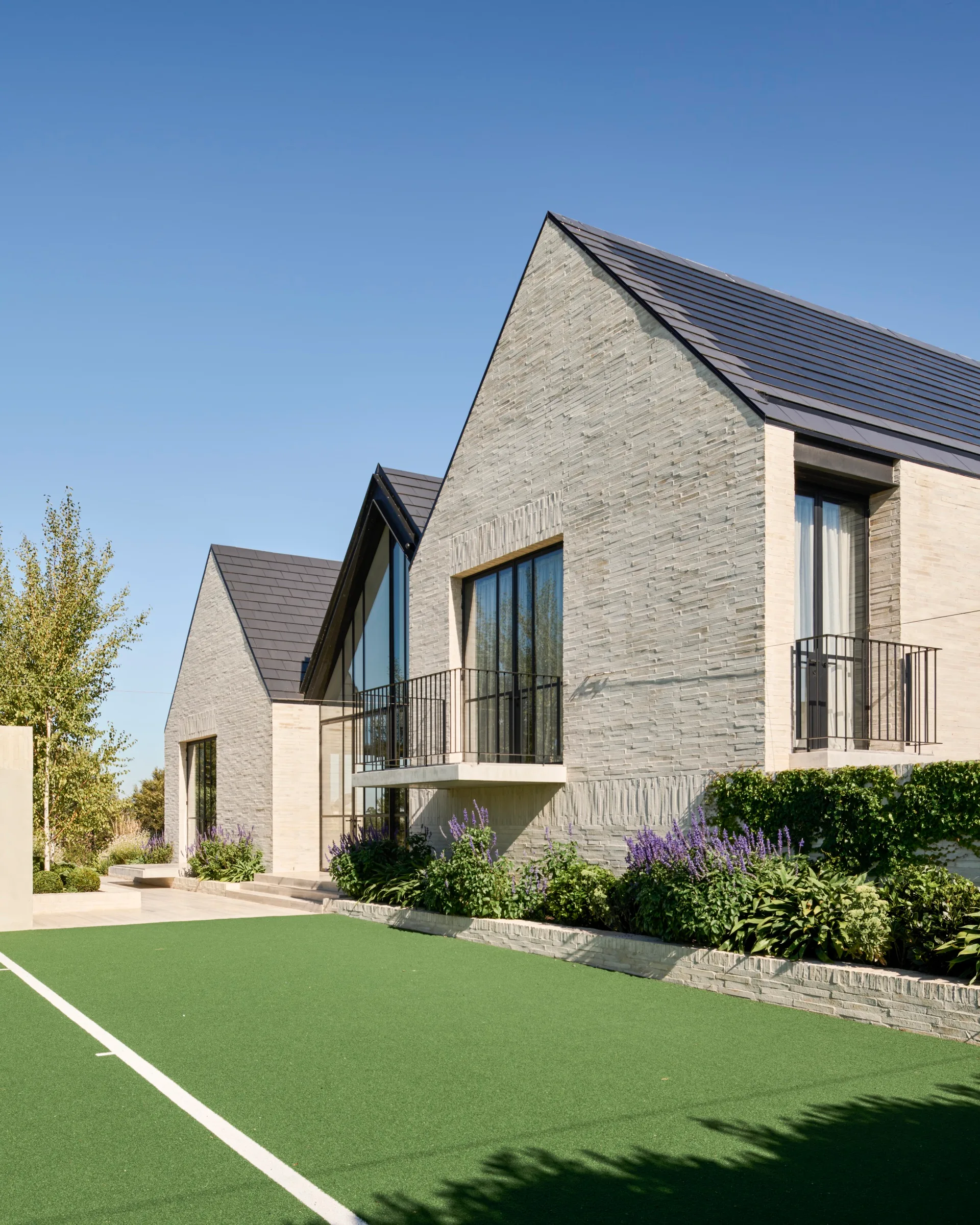 Modern house with gray brick exterior, gabled roof, large windows, and a manicured lawn under a clear blue sky.