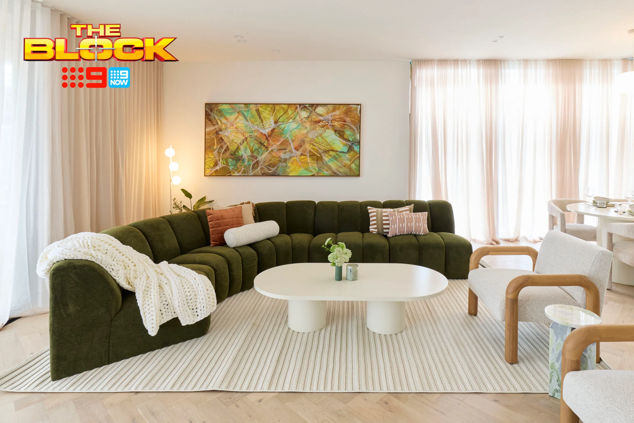 Living room with green sectional sofa, white table, abstract painting, and soft lighting from "The Block" show.