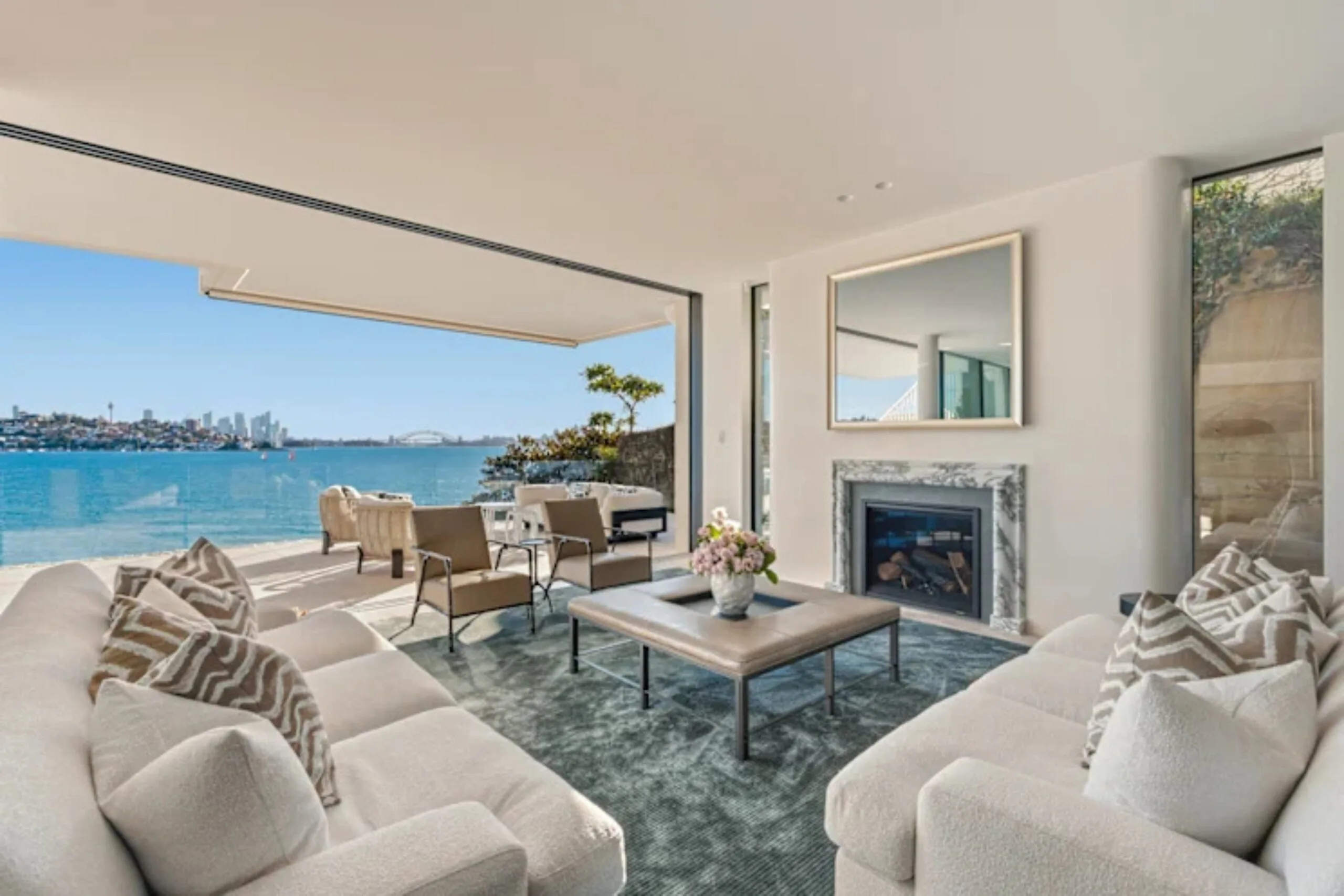 Luxurious living room with sofas, fireplace, and large glass doors opening to a stunning waterfront city view.