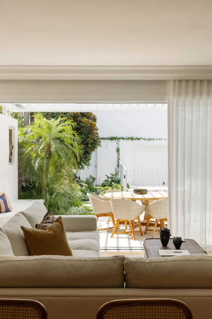 Bright living room with beige sofa, overlooking a patio with a dining table and lush greenery in the background.