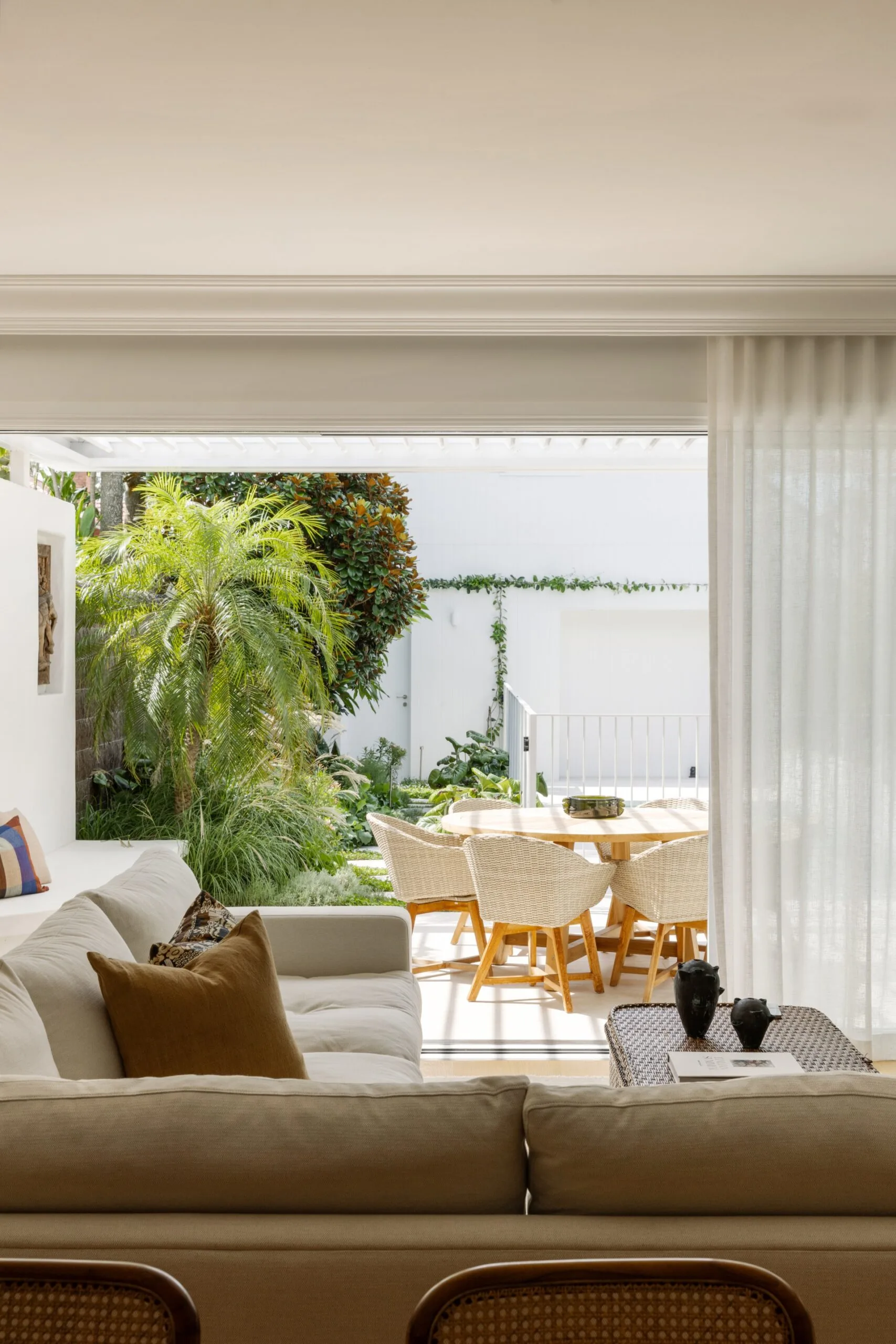 Bright living room with beige sofa, overlooking a patio with a dining table and lush greenery in the background.