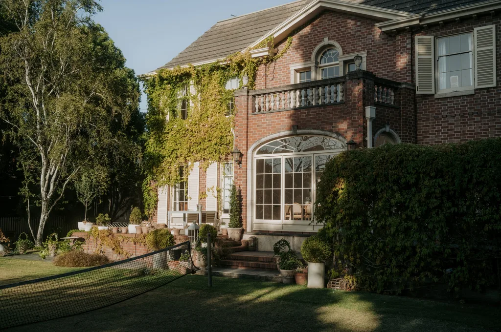 Red-brick house with ivy, large windows, balcony, and tennis net in sunlit garden.