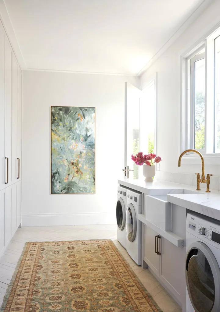 An all-white laundry with windows and a glass door opening outside, a tall painting hanging on the wall and a vintage-style runner on the tiled floor.