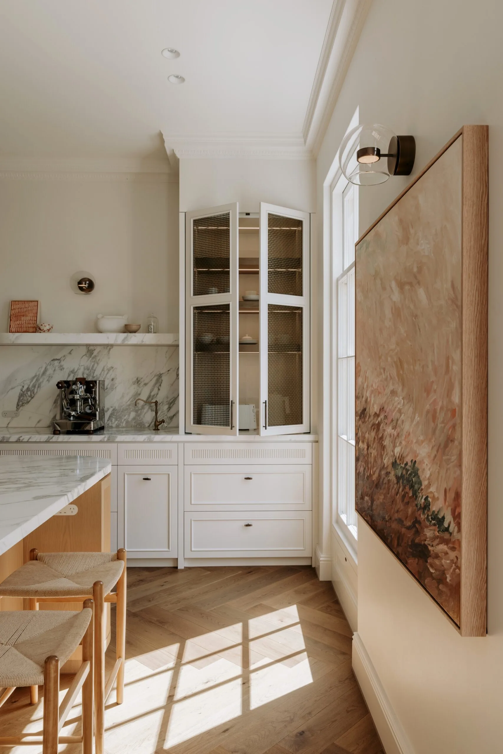 Modern kitchen corner with open cabinets, marble countertop, coffee machine, wooden stools, and large abstract painting.