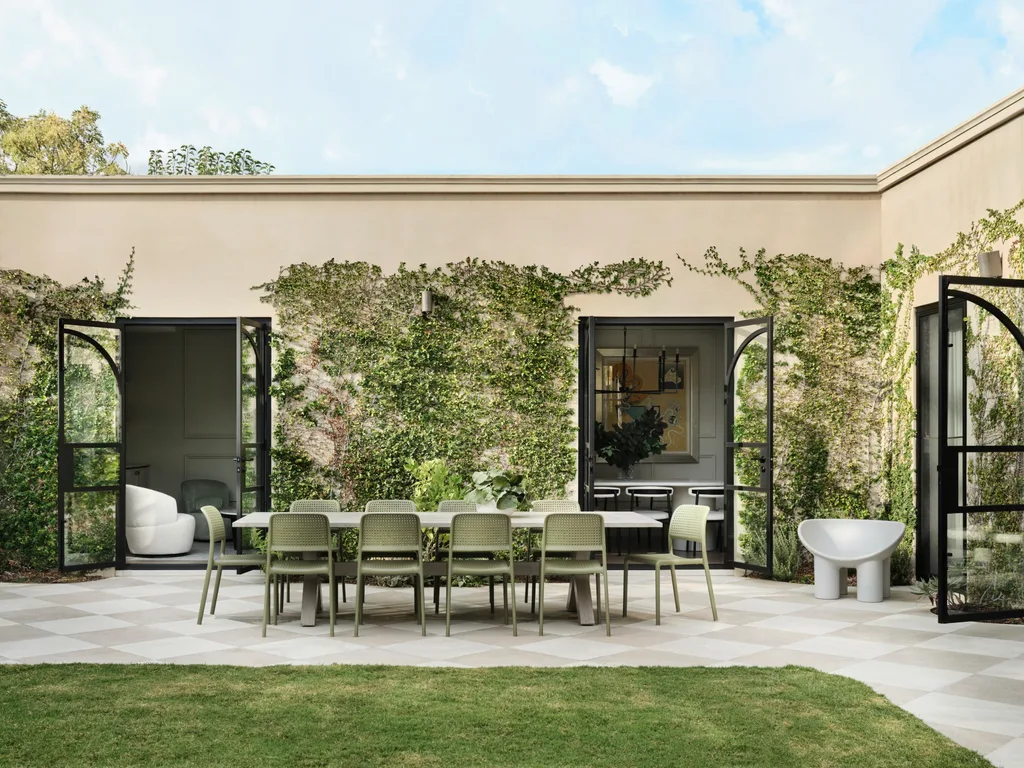 Outdoor patio with green chairs around a dining table, surrounded by ivy-covered walls and open glass doors.