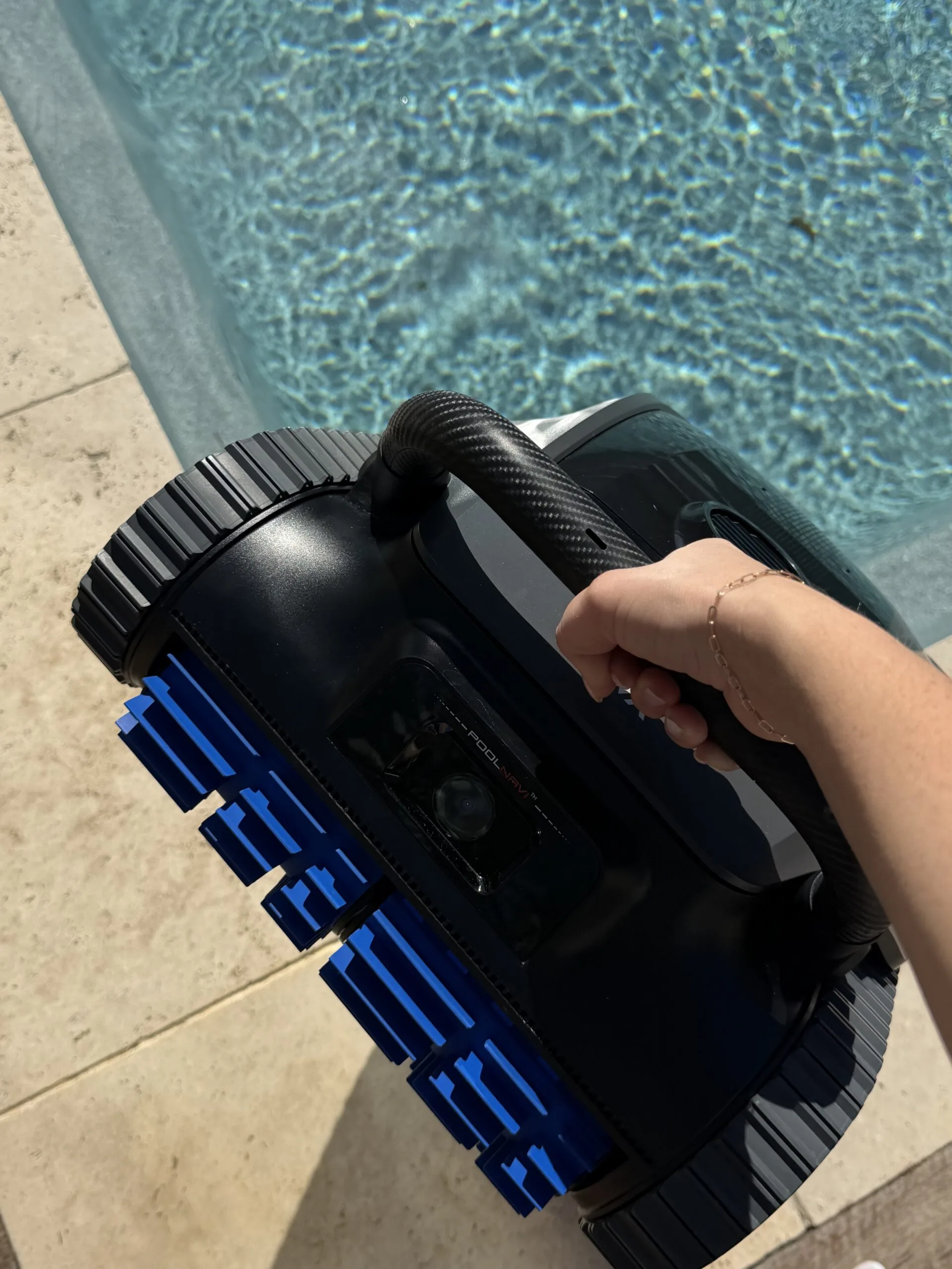 A hand holding a PoolLab robotic pool cleaner with blue brushes above a sunlit pool.