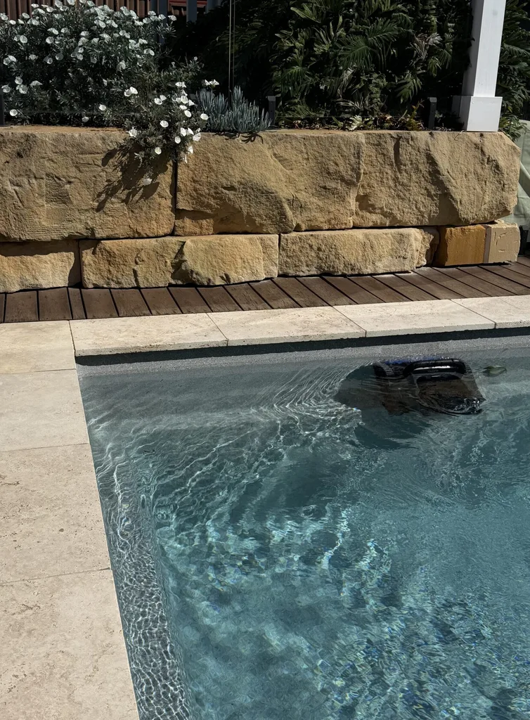 Sunny backyard with a clear pool, stone retaining wall, and lush plants set against a wooden deck.