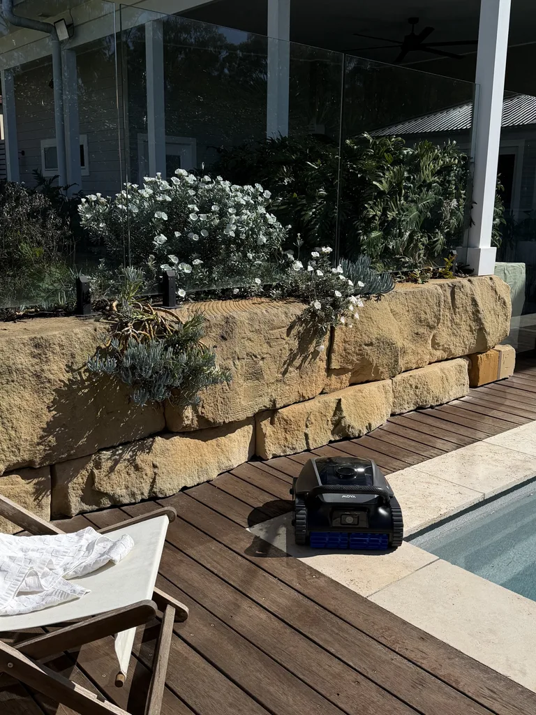 Poolside robot cleaner on wooden deck near stone wall and flowers, with a white chair in the foreground.